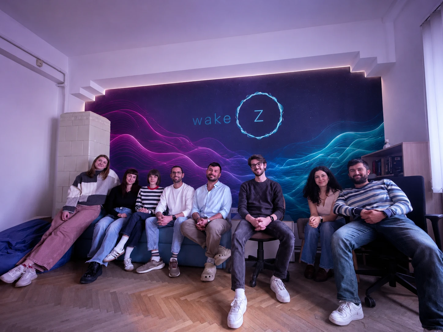 The WakeZ team, eight people sitting together in front of the WakeZ logo wall in the office.